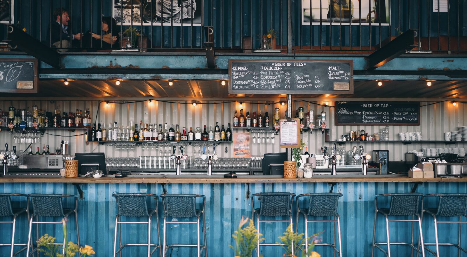 Industrial-style outdoor distillery bar with shelves of liquor bottles, beer taps, and chalkboard drink menus