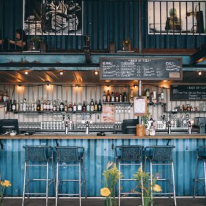 Industrial-style outdoor distillery bar with shelves of liquor bottles, beer taps, and chalkboard drink menus