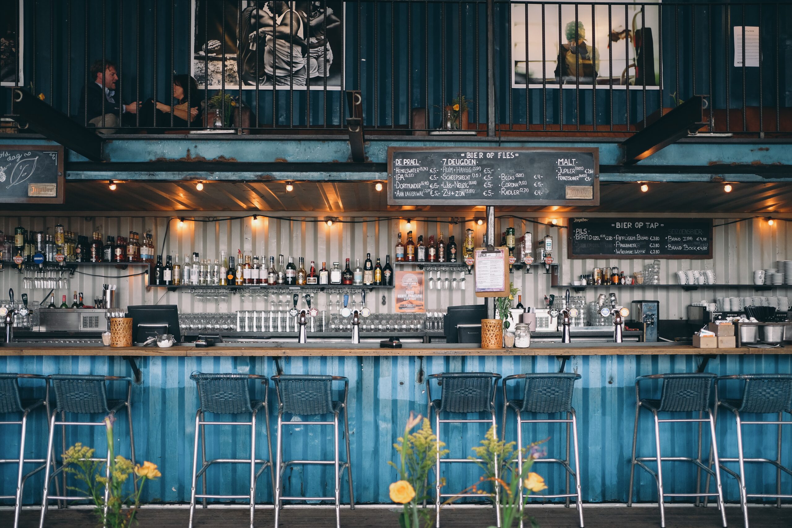 Industrial-style outdoor distillery bar with shelves of liquor bottles, beer taps, and chalkboard drink menus